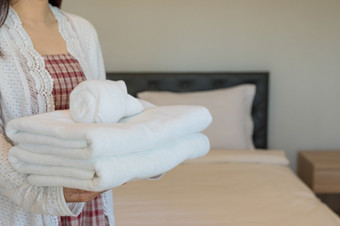 年轻的酒店女仆持有毛巾表和软毛巾的清洁房间的酒店有清洁白色毯子的床上与枕头的早....卧室软毛巾卧室