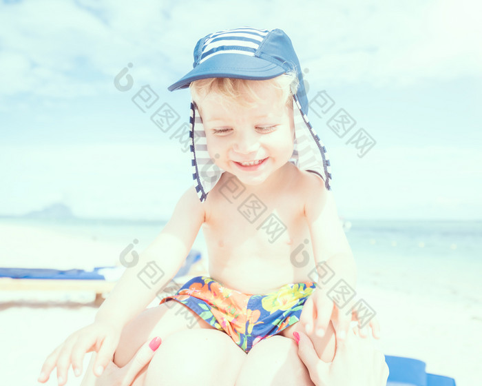 幸福婴儿男孩坐着妈妈她的腿的海滩使用分裂爽肤水