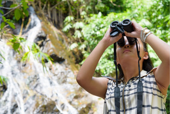 美丽的年轻的女孩徒步旅行使用双筒望远镜看为鸟热带森林附近的瀑布泰国