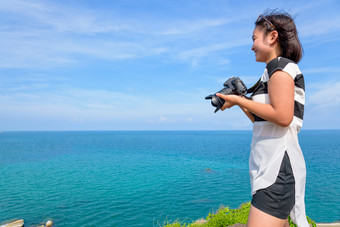 十几岁的女孩持有数码单反相机相机微笑幸福的的前的岛与的美丽的自然景观的蓝色的海和的夏天天空的背景十几岁的女孩持有相机高