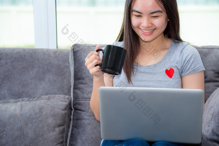 亚洲女人持有咖啡杯微笑和看在工作工作远程与移动PC的沙发的房子可爱的女孩青少年快乐使用互联网电脑沟通从首页女人持有咖啡杯在工作