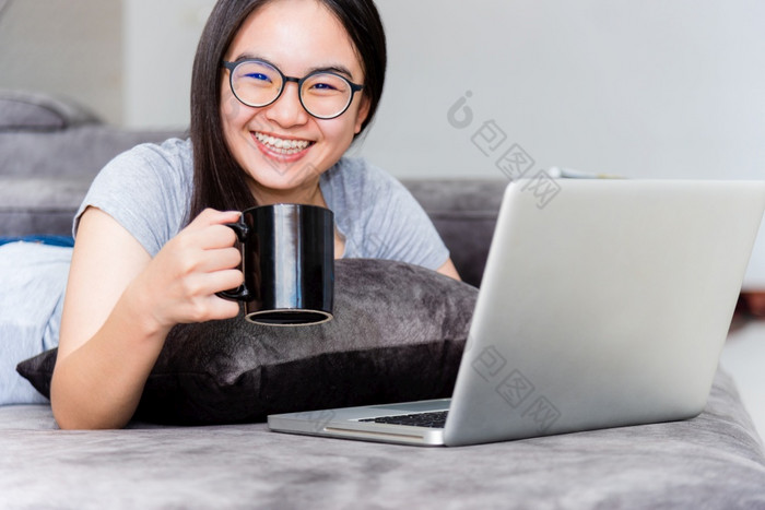 肖像美丽的亚洲年轻的女人持有咖啡杯微笑和看在工作可爱的女孩青少年快乐的沙发工作远程与移动PC和互联网电脑沟通从首页女人持有咖啡杯在工作