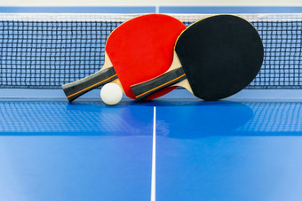 黑色的和红色的表格网球球拍和白色球的蓝色的平发出难闻的气味表格与网两个表格网球桨体育竞争设备室内活动和锻炼为背景概念黑色的和红色的表格网球桨与网