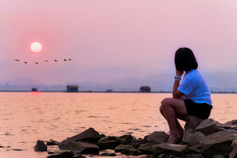 亚洲十几岁的女人坐孤独的独自一人的海滨看的美丽的自然景观太阳和群鸟飞行行日落的湖背景krasiao大坝suphan武里府泰国少年坐着独自一人海滨的日落
