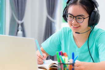 年轻的亚洲女人学生与耳机和眼镜写作的书看视频会议移动PC电脑女孩快乐取的大学互联网距离学习在线类亚洲女人学生快乐类在线学习