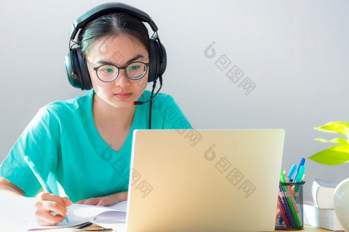 亚洲年轻的女人学生与眼镜耳机研究严重的写作请注意书看视频会议移动PC电脑大学类在线互联网学习距离教育首页亚洲女人学生快乐类在线学习