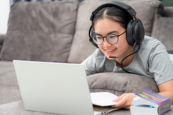 亚洲年轻的女人学生与耳机说谎的沙发微笑看移动PC在线学习从学校女孩快乐大学互联网距离研究在线工作电脑从首页距离学习在线从首页