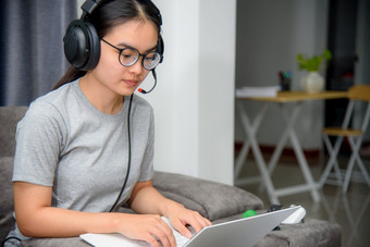 亚洲年轻的女人学生与耳机坐着的沙发看移动PC快乐研究在线类大学学习互联网教育十几岁的女孩工作距离移动PC电脑从首页距离学习在线从首页