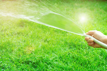 老女人手持有橡胶水软管和使用手指关闭结束橡胶水软管使水喷雾与阳光和绿色草场背景