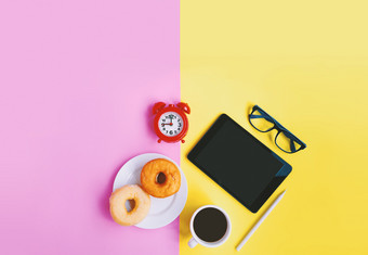 现代首页工作场所红色的报警时钟杯咖啡甜甜圈眼镜和平板电脑黄色的粉红色的柔和的背景与复制空间
