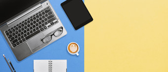 工作空间的现代首页办公室桌面与移动PC平板电脑和热咖啡泡沫喝色彩斑斓的黄色的蓝色的背景平铺桌面高角视图与复制空间