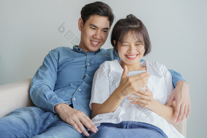 快乐年轻的爱亚洲夫妇坐着沙发上首页看移动电话年轻的亚洲人是使用智能手机