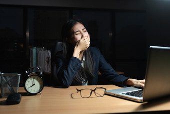 亚洲业务女人坐着打呵欠与手覆盖她的口移动PC的桌子上而的年轻的女人作品加班的办公室晚上