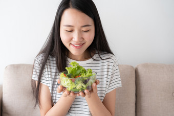 亚洲女人持有<strong>玻璃碗</strong>完整的沙拉<strong>蔬菜</strong>绿色而坐着的沙发首页健康的和生活方式概念