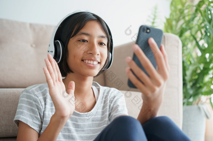 亚洲小女孩视频调用与耳机的智能手机和挥舞着问候而坐着的生活房间首页概念放松和生活方式首页