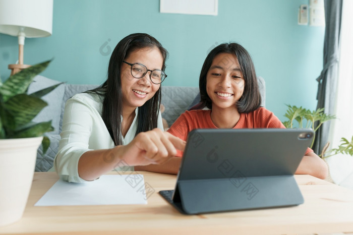 亚洲家庭与女儿家庭作业使用平板电脑与妈妈。帮助快乐微笑亚洲孩子而坐着的生活房间首页早....概念在线学习首页