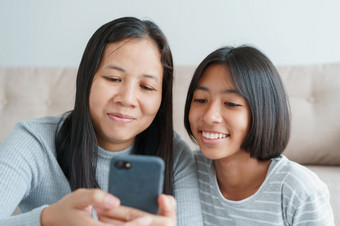 家庭亚洲妈妈。和女儿使用智能手机在一起首页快乐女孩和妈妈而坐着的生活房间在一起特写镜头脸孩子和妈妈