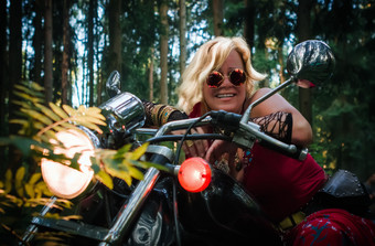 有吸引力的成熟的金发女郎女人骑摩托车的人轮黑暗眼镜服装嬉皮风格坐着摩托车与的灯的森林成熟的女人骑摩托车的人摩托车