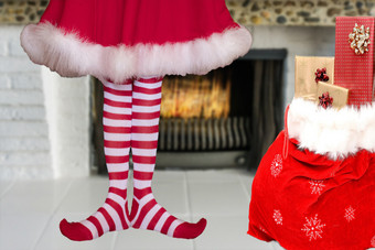 可爱的可爱的小圣诞节精灵女孩与尖尖的脚穿条纹精灵长袜和红色的衣服站下一个圣诞老人老人袋完整的礼物前面燃烧壁炉