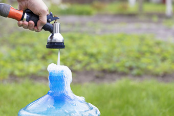 的园丁持有灌溉喷水灭火系统和收集清洁水蓝色的瓶对模糊明亮的绿色草的园丁持有灌溉喷水灭火系统和收集清洁水