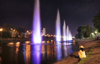 女孩黄色的衣服坐在的银行的河的草和看起来的美丽的明亮的和高喷泉的rusanovskaya通道的第聂伯河路堤城市景观基辅美丽的明亮的和高喷泉的rusanovskiy通道的晚上基辅