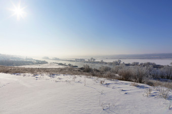 农村冬天景观的太阳rsquo射线阴霾蓝色的天空谎言绕组河和白雪覆盖的字段灌木和树明亮的太阳与阴霾的蓝色的天空照亮的河和白雪覆盖的字段的冷淡的早....