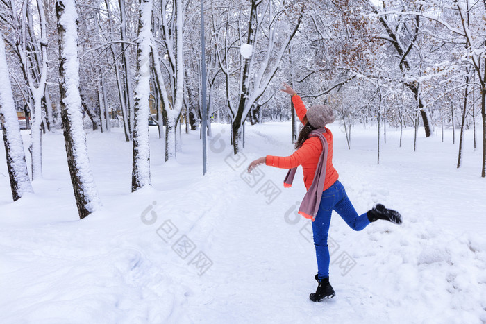 年轻的美丽的女人明亮的珊瑚夹克和蓝色的牛仔裤冬天走沿着的小巷的白雪覆盖的童话语言公园和抛出大雪球年轻的美丽的女人走冬天沿着小巷白雪覆盖的令人难以置信的城市公园