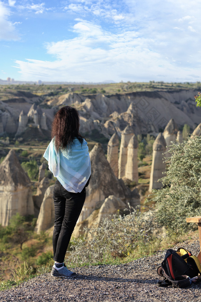 年轻的女孩绿松石围巾她的肩膀站的边缘悬崖和看起来的山峡谷和蓝色的天空卡帕多西亚年轻的女孩站的边缘悬崖和看起来的地平线的山山谷卡帕多西亚和的蓝色的天空