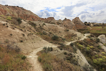 山小道蜿蜒沿着路在灌木之间的巨大的老锥形和饱经风霜的岩石的红色的谷卡帕多西亚下多云的春天天空绕组山小道通过之间的巨大的和老石头的不过红色的谷