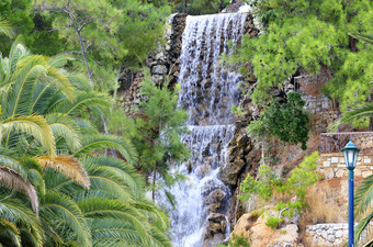 大瀑布从春天氡水流下来大巨石的脚的山在的绿色地中海松树和棕榈树的公园洛特拉基希腊大瀑布与氡水在巨石的脚山洛特拉基希腊