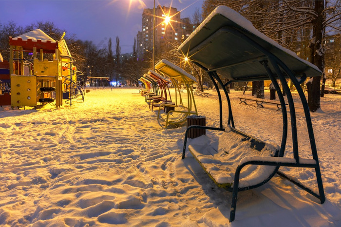 空操场上与摆动木长椅与金属栏杆的雪对的蓝色的《暮光之城》背景冬天晚上城市公园和照亮的温暖的光街灯木长椅和操场上的冬天城市晚上公园是覆盖与雪对的背景蓝色的《暮光之城》