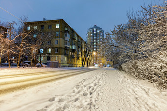 冬天城市景观与白雪覆盖的路沿着城市街与住宅建筑和树的雪与城市晚上照明对的背景蓝色的《暮光之城》白雪覆盖的路沿着城市街与树的雪和城市晚上照明对的背景蓝色的《暮光之城》