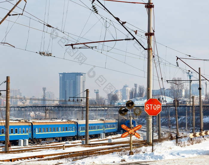 红色的停止标志铁路穿越对的背景铁路跟踪蓝色的火车车厢和城市景观的入口冬天城市红色的停止标志的铁路穿越对的背景的铁路站和建筑的冬天城市