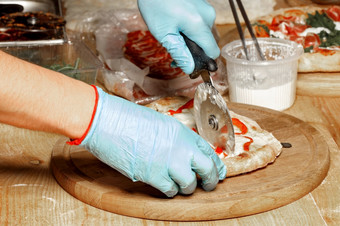 的手老板高加索人男人。蓝色的乳胶手套是切割的煮熟的披萨木厨房董事会特写镜头的老板手蓝色的乳胶手套减少的煮熟的披萨与轮刀