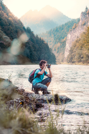 年轻的旅游与背包看起来通过双筒望远镜山山峰站岩石在河男孩花假期山流浪的与背包穿体育夏天衣服