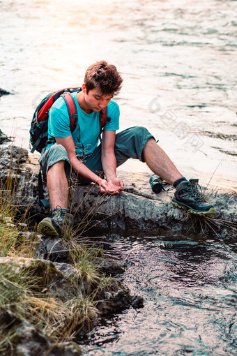 年轻的男孩需要纯水从河和持有的手坐着岩石在的河休息在徒步旅行花假期流浪的与背包穿体育运动夏天衣服