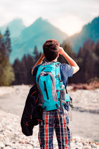 年轻的流浪者与背包看起来通过双筒望远镜山山峰站在河男孩花假期山流浪的与背包穿体育夏天衣服