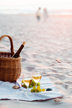两个酒眼镜与白色酒站沙子海滩旁边葡萄和柳条篮子与瓶酒年轻的夫妇站的海的背景