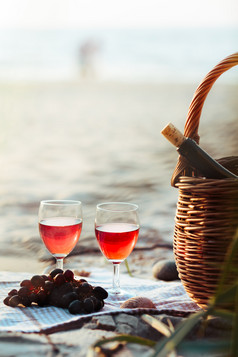 两个酒眼镜与红色的酒站海滩旁边葡萄和柳条篮子与瓶酒年轻的夫妇站的海的背景