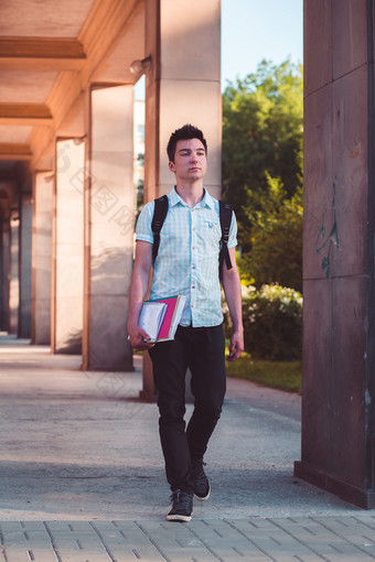 学生持有笔记本和携带背包走的前面大学建筑年轻的男孩穿蓝色的衬衫和黑暗牛仔裤