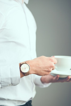 年轻的男人。穿银手表和白色平原衬衫持有杯咖啡站前面平原灰色墙
