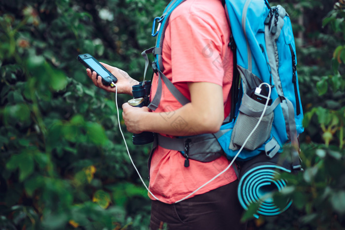 年轻的男人。使用在线地图在的徒步旅行使用聪明的电话充电移动电话在的旅行持有背包
