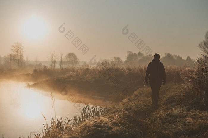 女人走通过草地池塘的多雾的早....太阳不断上升的以上场和池塘淹没了与雾的早....真正的人真实的的情况下