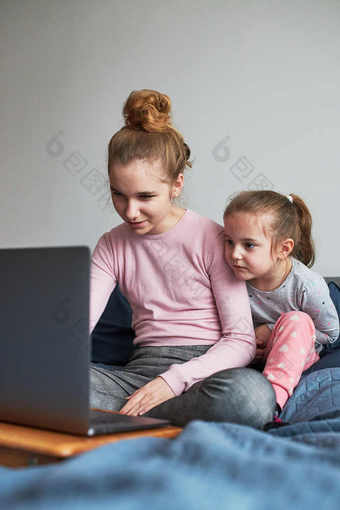 十几岁的女孩和她的年轻的妹妹玩学习移动PC支出时间在一起首页在新冠病毒检疫女孩坐着床上前面电脑
