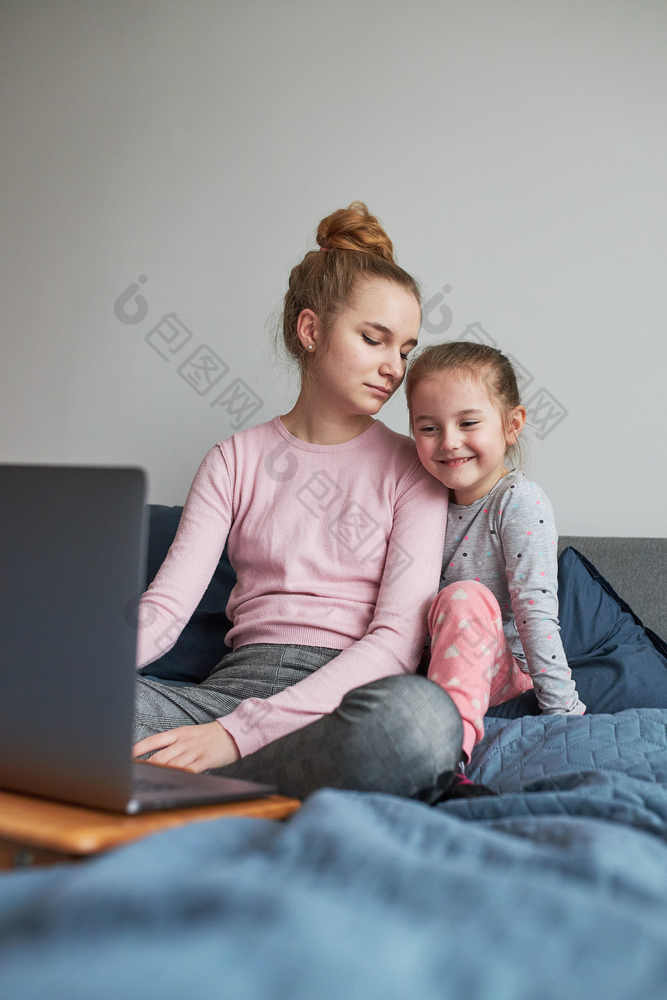 十几岁的女孩和她的年轻的妹妹玩学习移动PC支出时间在一起首页在新冠病毒检疫女孩坐着床上前面电脑