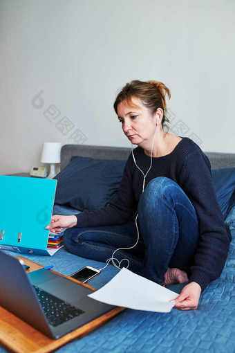 女人工作做她的工作远程在视频闲谈，聊天调用流在线课程网络研讨会移动PC从首页女人坐着床上前面电脑看屏幕使用耳机和智能手机