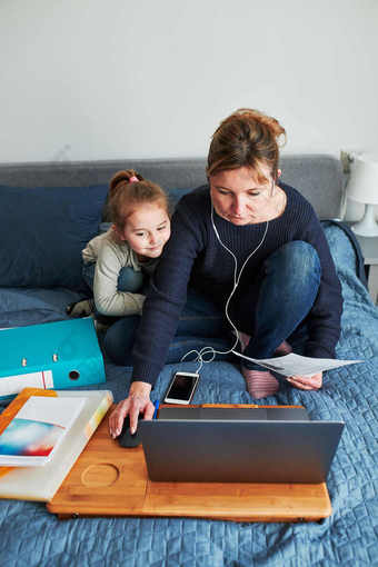 女人妈妈。工作做她的工作远程在视频闲谈，聊天调用流在线课程网络研讨会移动PC从首页而她的女儿玩周围在新冠病毒检疫女人坐着床上前面电脑看屏幕使用耳机和智能手机