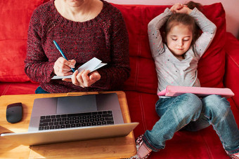 女人妈妈。工作做她的工作远程在视频闲谈，聊天调用流在线课程网络研讨会移动PC从首页在新冠病毒检疫而她的女儿看视频平板电脑女人坐着床上前面电脑看屏幕