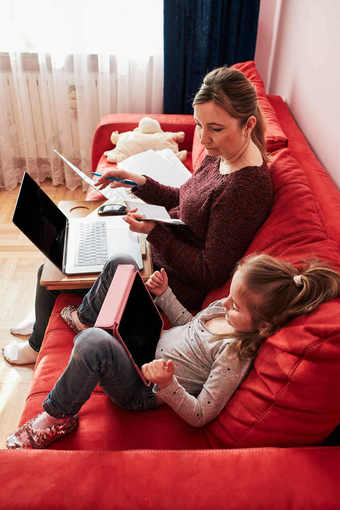 女人妈妈。工作做她的工作远程在视频闲谈，聊天调用流在线课程网络研讨会移动PC从首页在新冠病毒检疫而她的女儿看视频平板电脑女人坐着床上前面电脑看屏幕