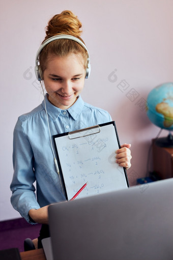 年轻的女人学生显示家庭作业有类学习在线看教训远程听教授会说话的与同学们视频调用从首页在检疫年轻的女孩使用移动PC耳机书手册坐着桌子上